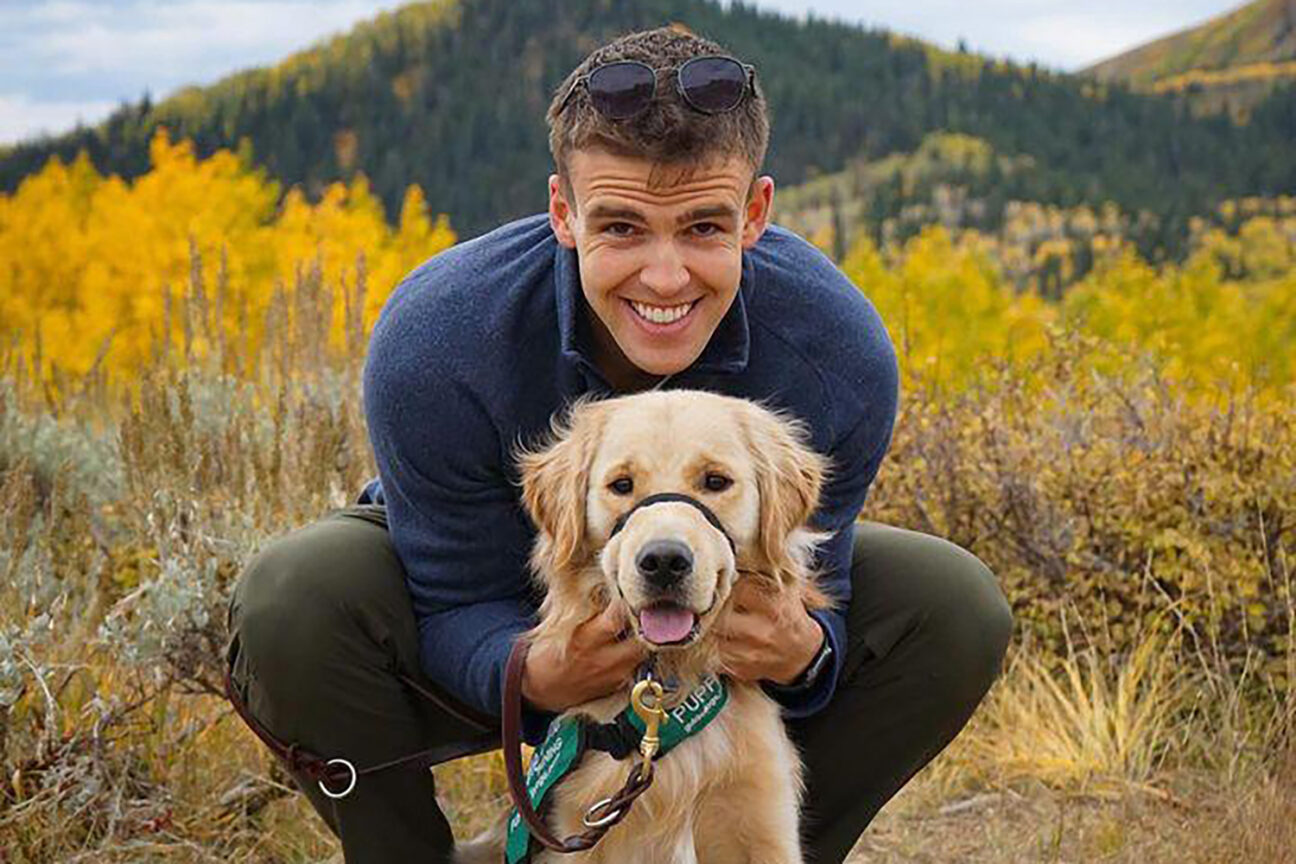 A puppy raiser and his guide dog puppy posing for a photo with a backdrop of mountains and fall colors.