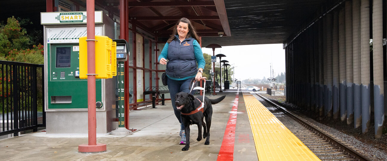 A guide dog mobility instructor walks with a guide dog on a train platform.