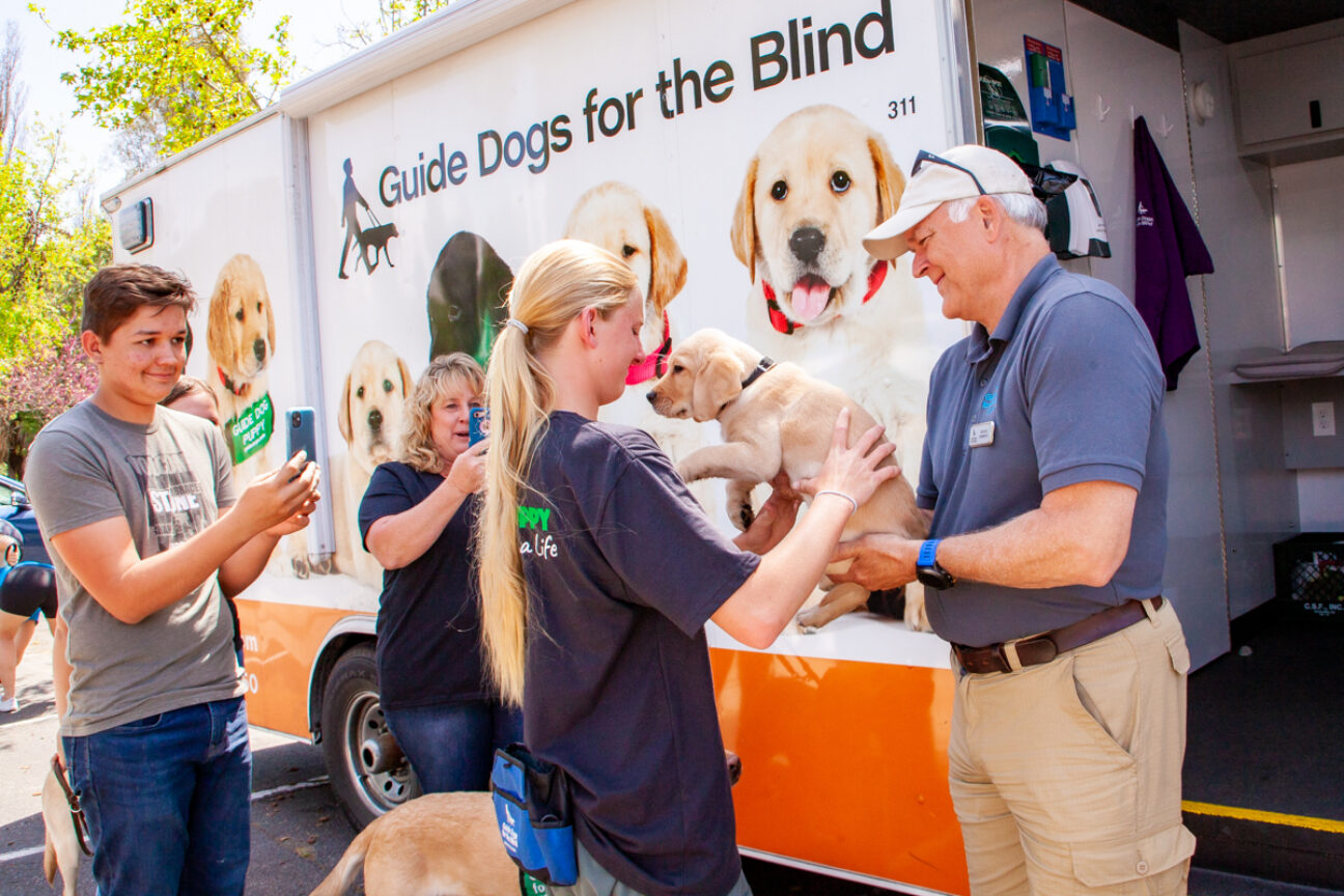 A young girl being handed a young yellow Lab puppy as friends look on.