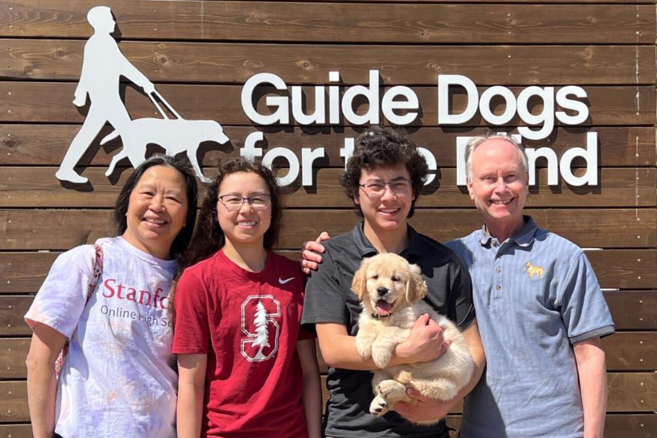 A family pose for a photo with a Golden Retriever puppy in front of a Guide Dogs for the Blind sign.