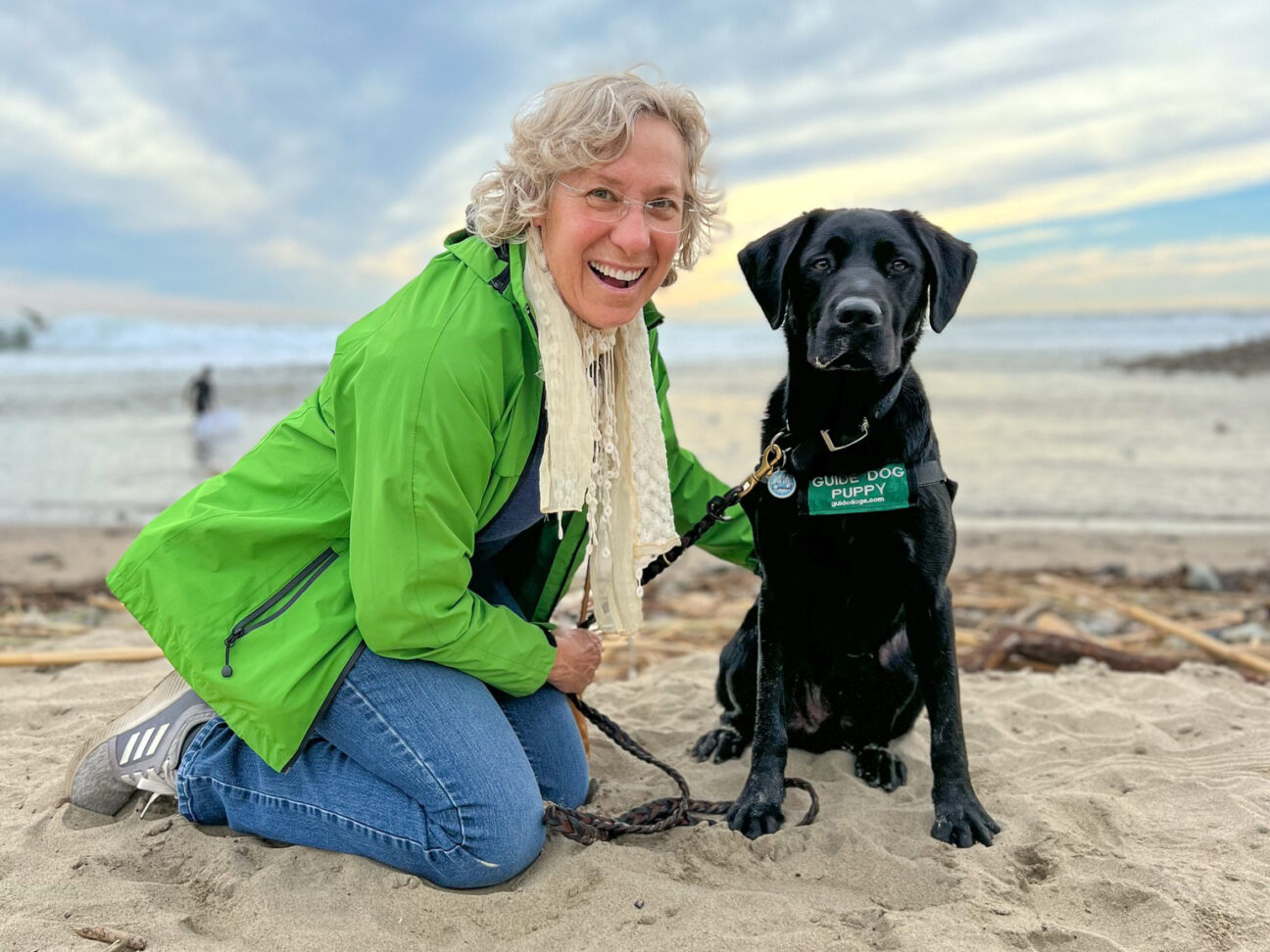 A woman on a beach with a black Lab guide dog puppy.