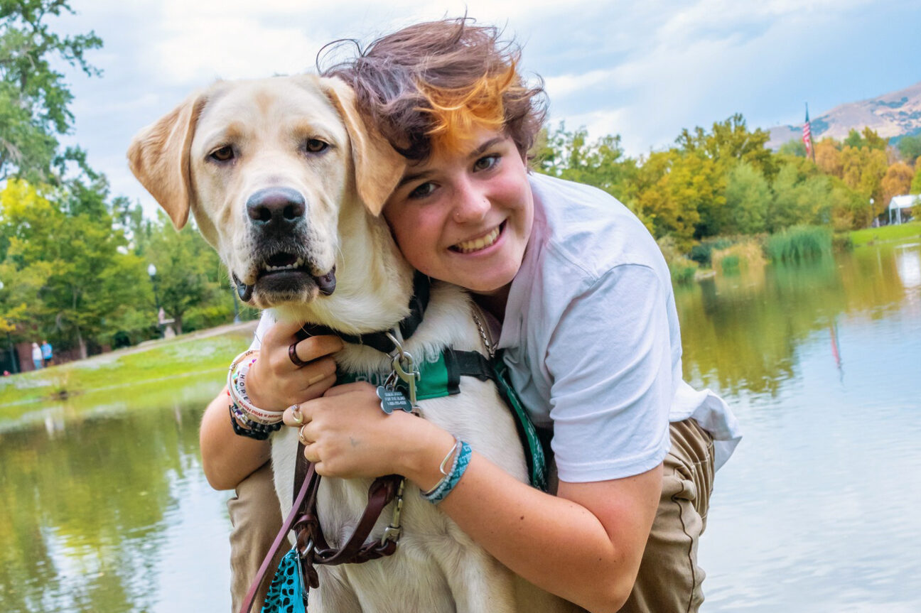 In a scenic park with a lake in the background, a puppy raiser hugs their guide dog puppy.