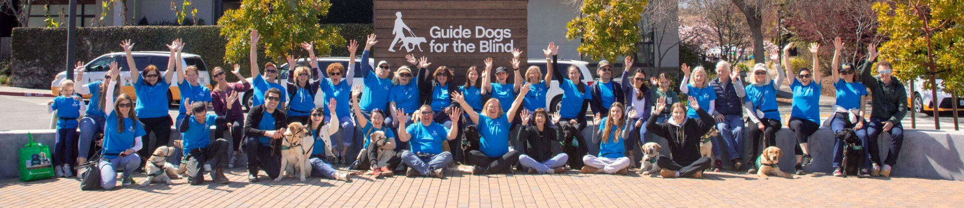 A large group of people and puppies pose for a photo in front of a sign with the Guide Dogs for the Blind logo.