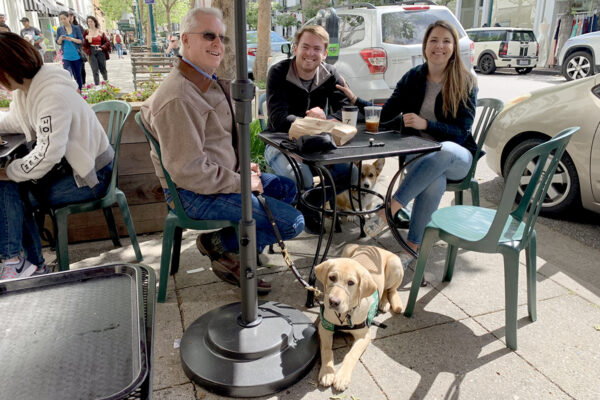 A guide dog puppy settled beneath an outdoor cafe table where three people are seated.