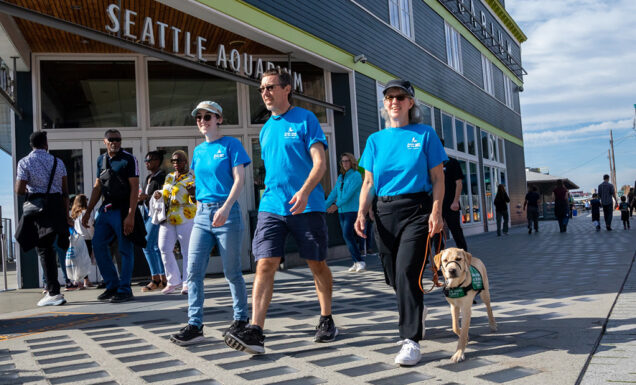 Three people walk with a guide dog puppy on the sidewalk in front of the Seattle Aquarium.
