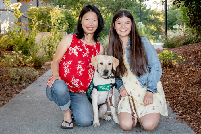 Mother and daughter kneel next to a yellow Lab guide dog puppy.