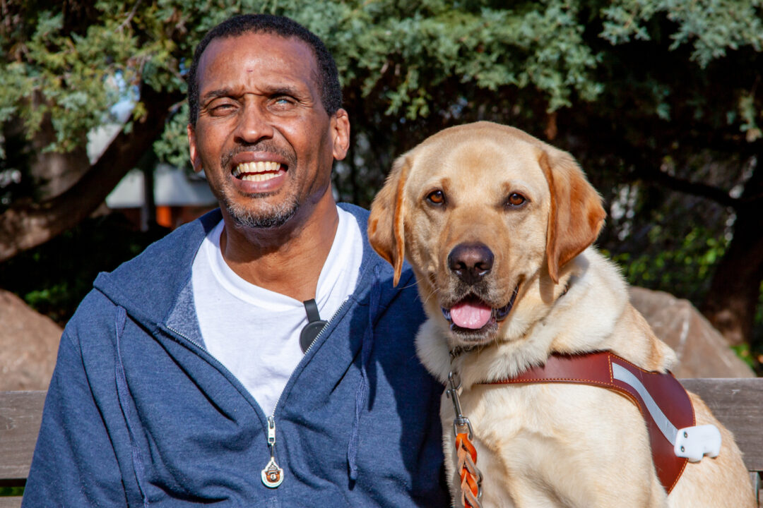 A smiling man sits next to a smiling guide dog.