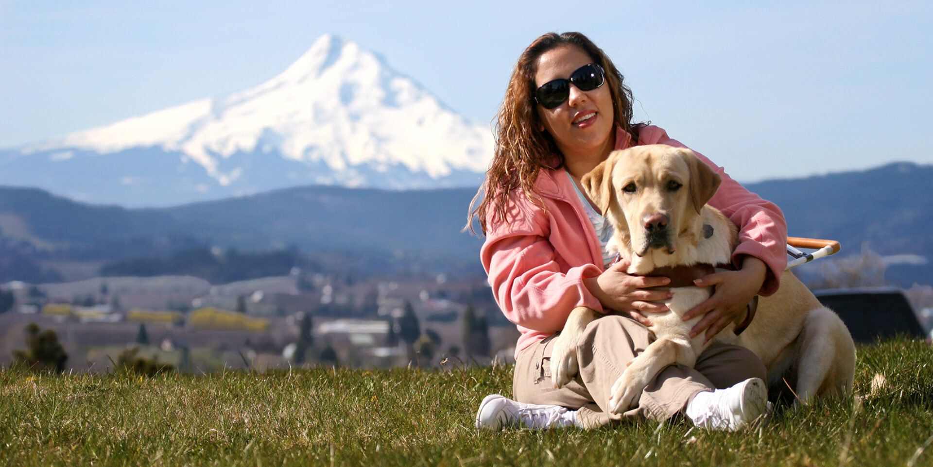 A woman sits on the grass with her guide dog; a panorama view of Mt. Hood is in the background.