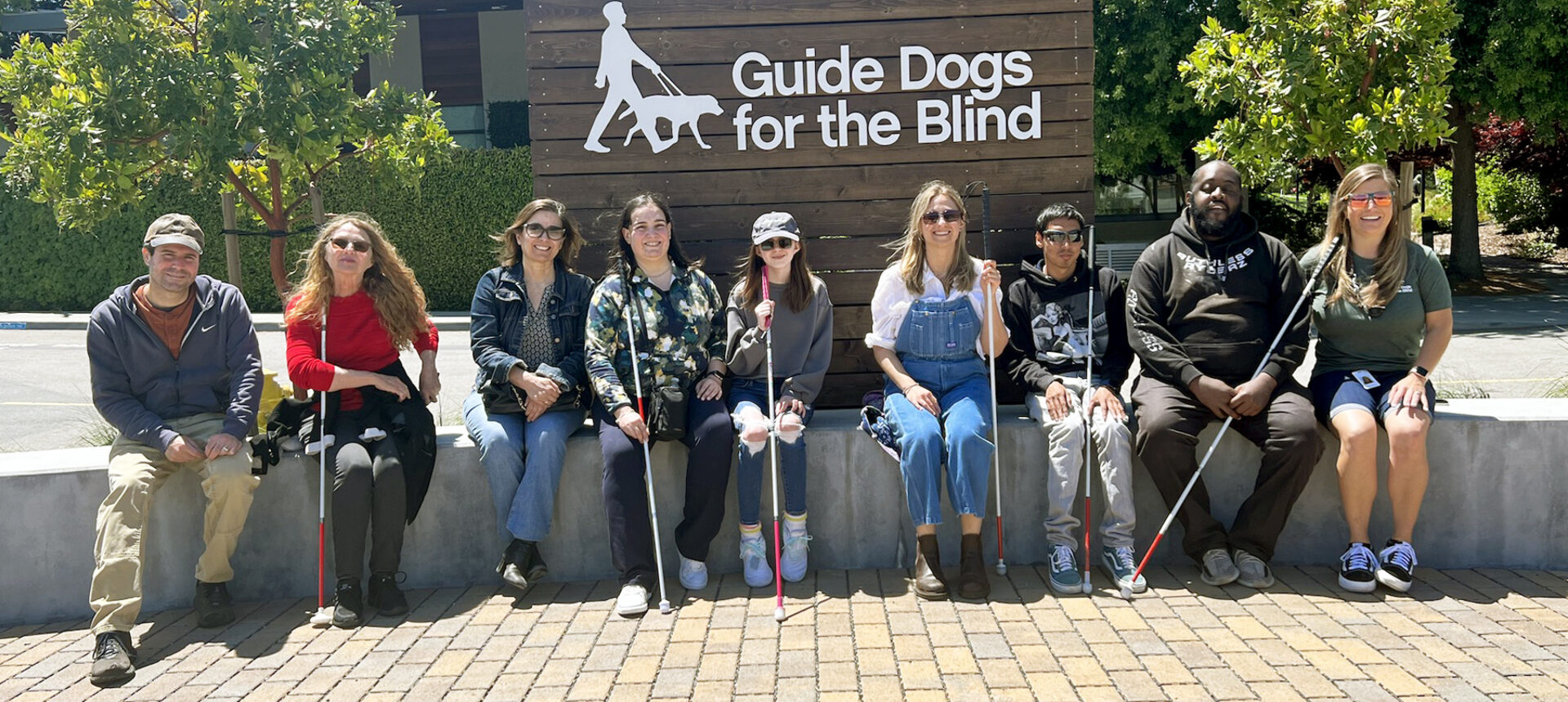 A group of students with white canes and their teachers seated in front of a Guide Dogs for the Blind logo sign.