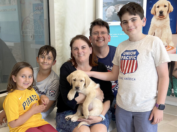 A family of five pose for a photo with a baby yellow Lab puppy.
