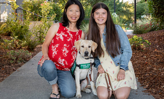 A mother and daughter kneel next to a yellow Lab guide dog puppy.