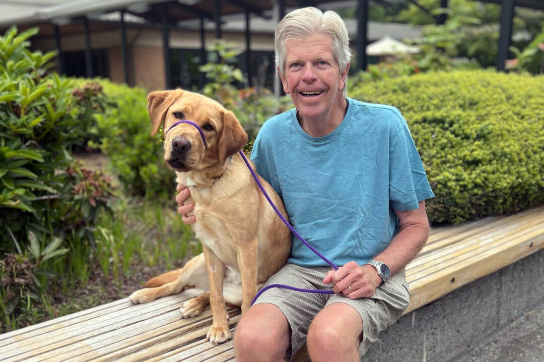 A smiling man sits on a bench next to a yellow Lab.