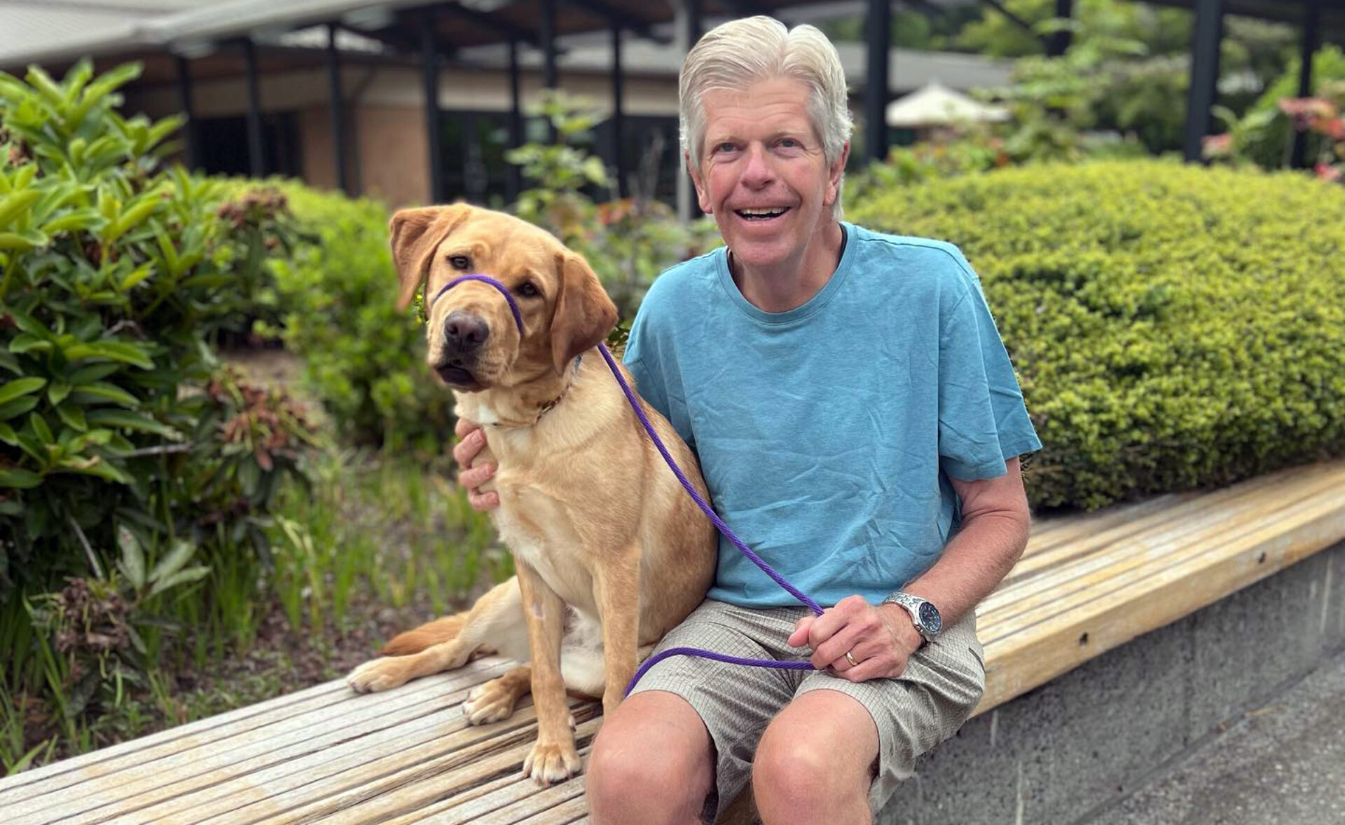 A smiling man sits on a bench next to a yellow Lab.
