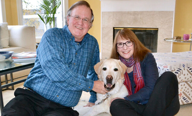 A happy couple snuggle with a smiling yellow Lab intheir home.
