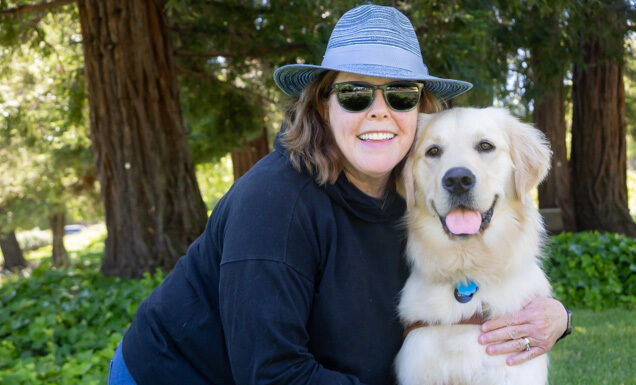A woman in a blue sun hat embraces a smiling Golden Retriever guide dog.