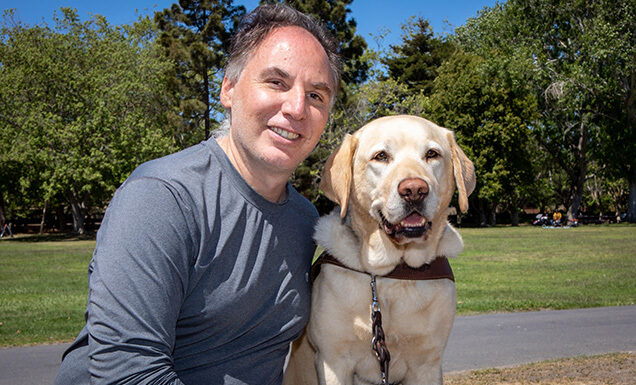 A man sits next to a yellow Lab guide dog.