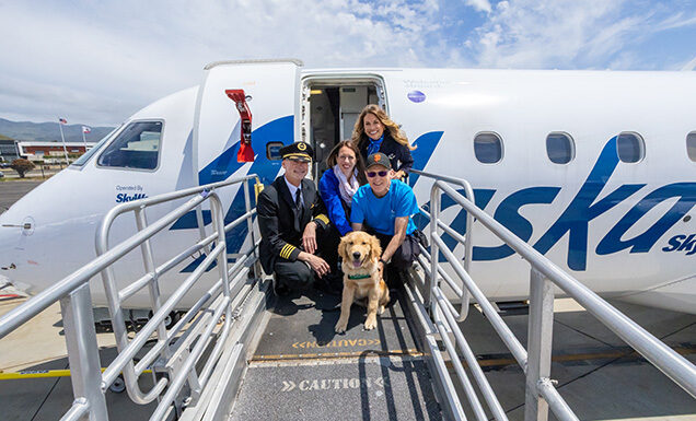 Two Alaska Airlines employees pose outside a plane door with two people and a guide dog puppy.