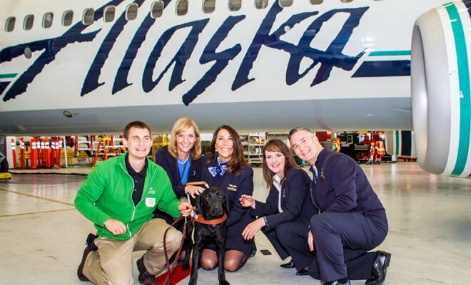 A group of airline personnel pose in front of an Alaska Airlines plane with a man and his guide dog.