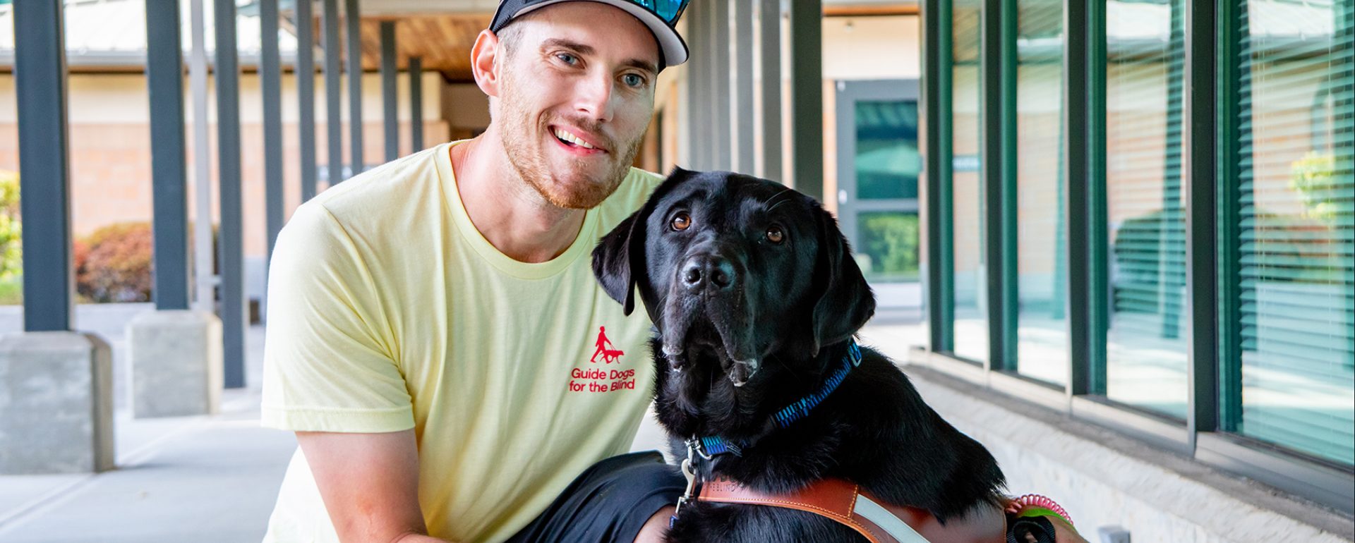 A person kneeling next to a black Lab guide dog.