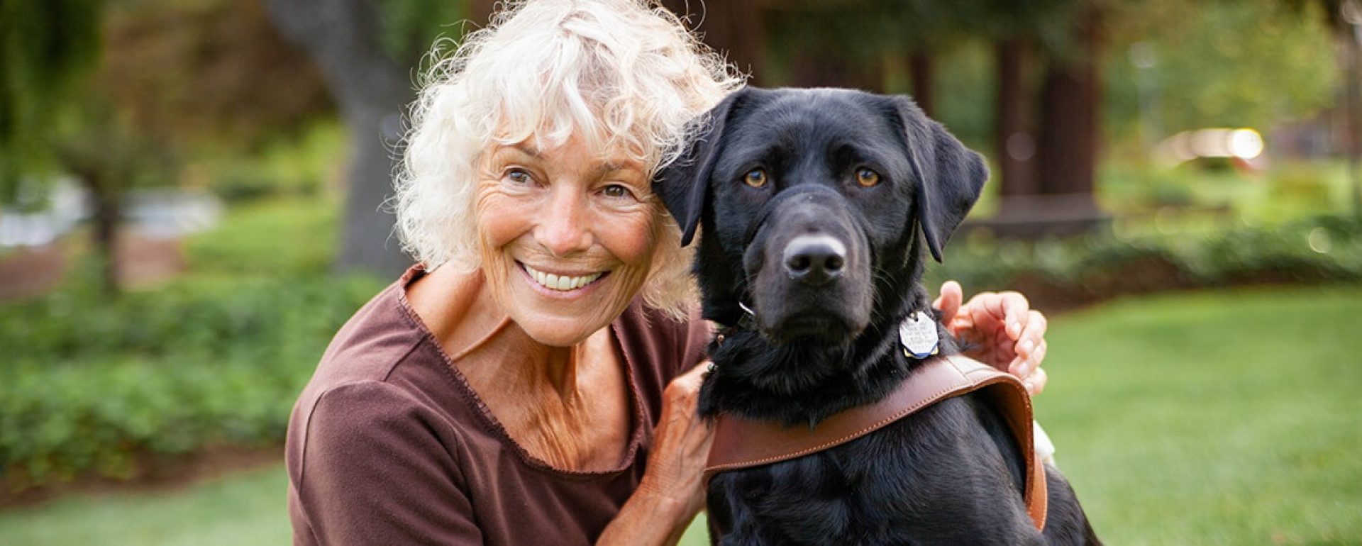 A woman sitting on a lawn hugging her black Lab guide dog.
