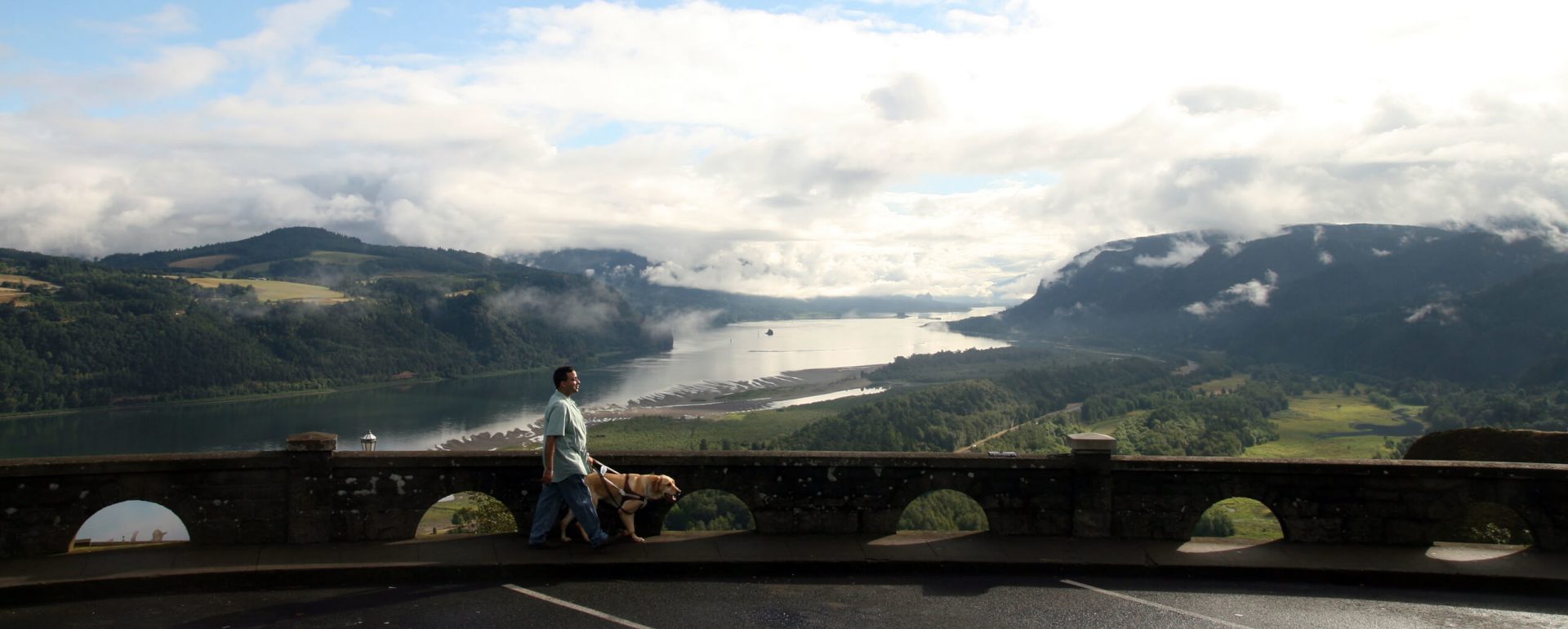 A GDB graduate walks with his yellow Lab guide dog along Crowne Point in Oregon with the beautiful Columbia Gorge in the background