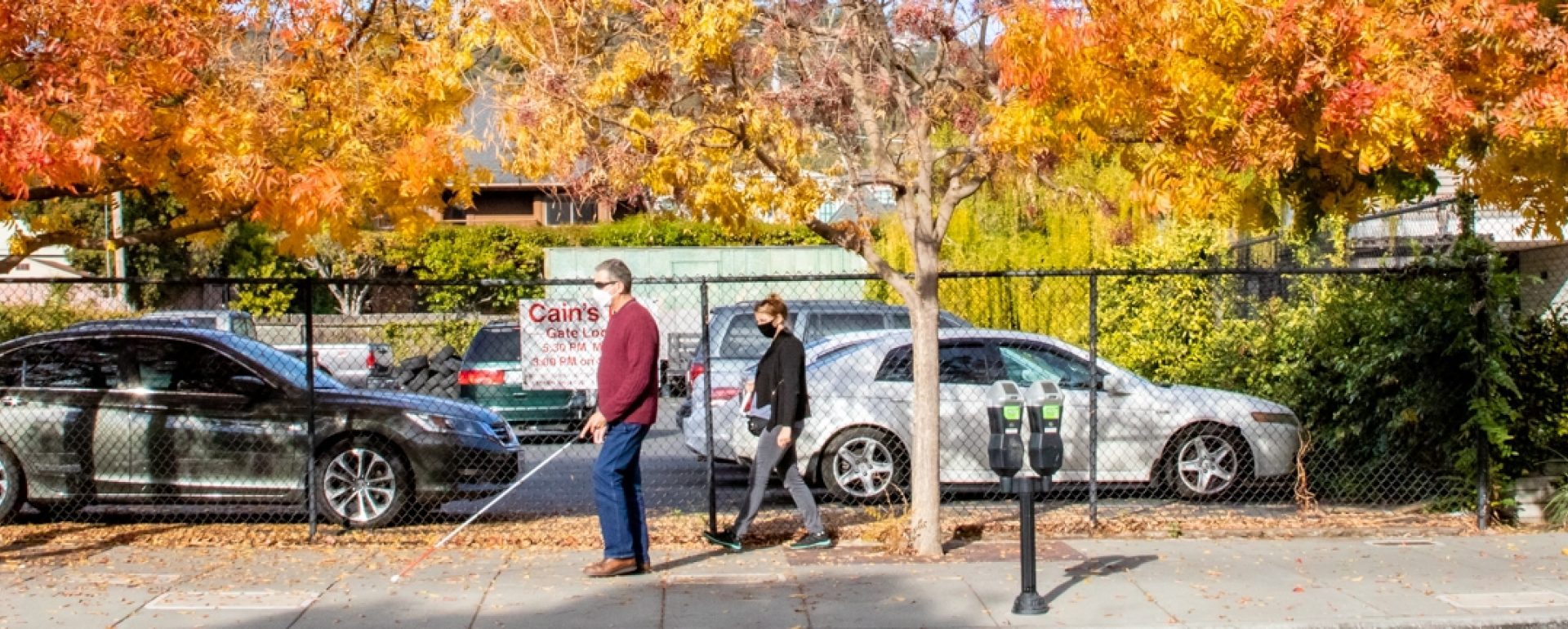 A person walks along a sidewalk using a white cane, followed by a mobility instructor.