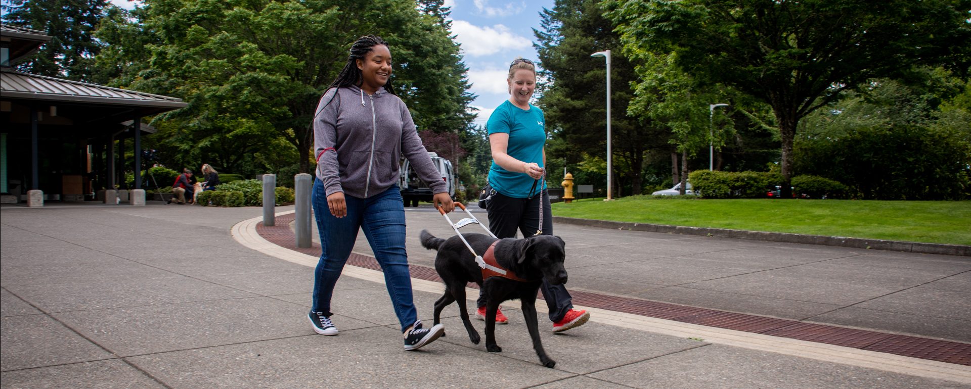 A young woman walks with a black Lab guide dog, accompanied by a guide dog mobility instructor. 