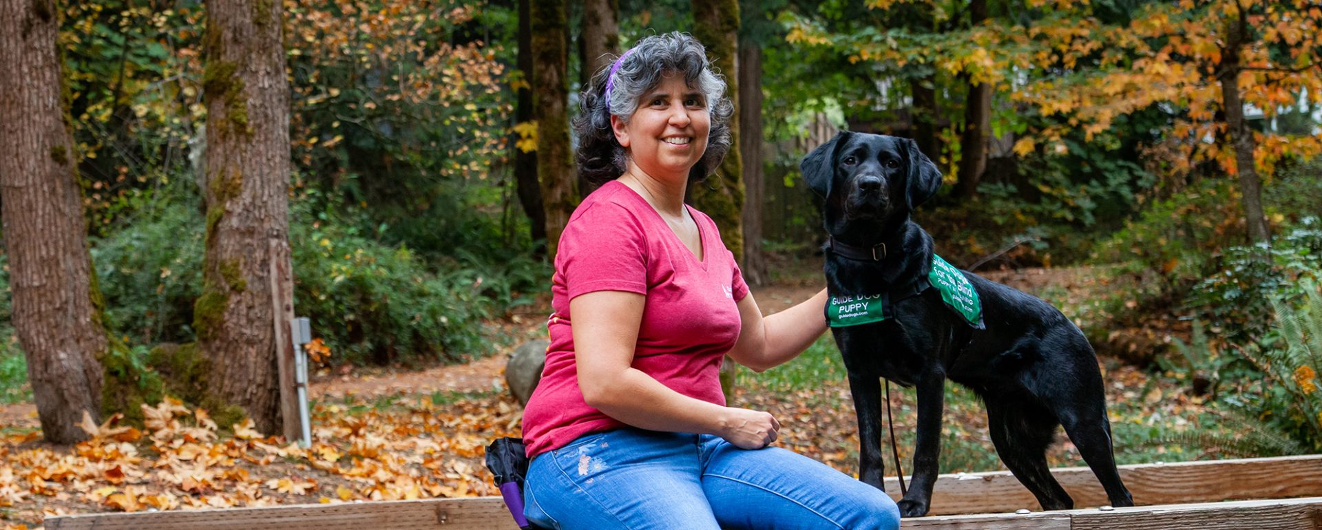 A woman sits next to a black Lab guide dog puppy in a wooded park.