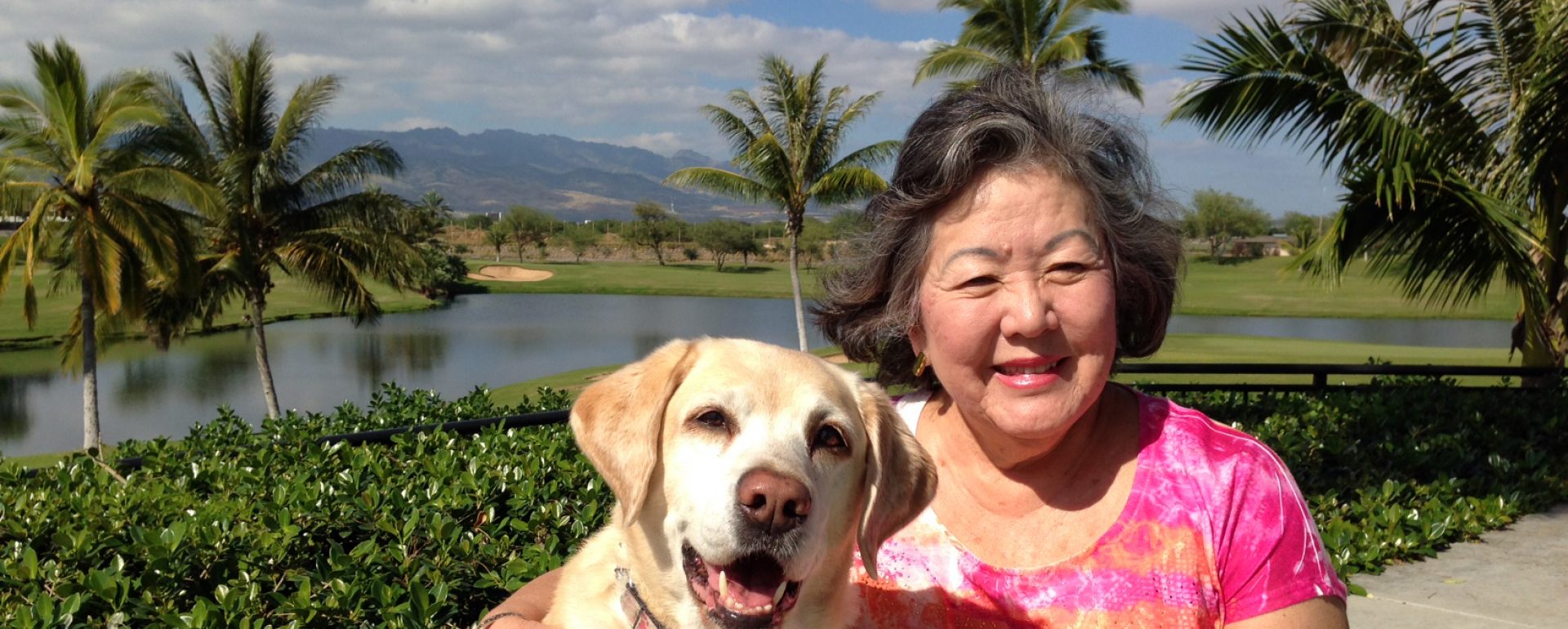 A female GDB graduate smiles next to her yellow Lab guide dog with palm trees in the background