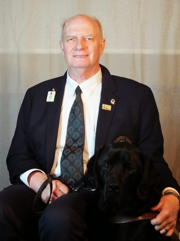 A portrait photograph of a man wearing a suit and sitting next to his guide dog.