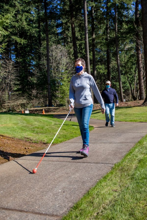 GDB Client Sherry Brackett navigates a wooded trail with a white cane. O&M Manager Marc Gillard follows behind her.