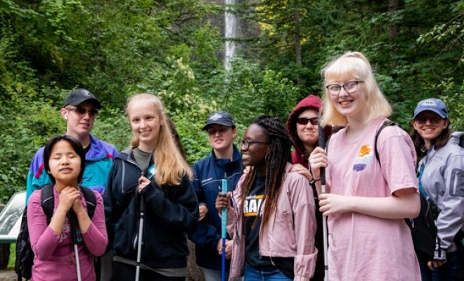 A group of youth campers shown on a hike in front of a waterfall.