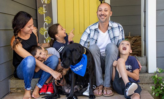 A family sits and laughs on their front porch with a black Lab GDB breeder dog.