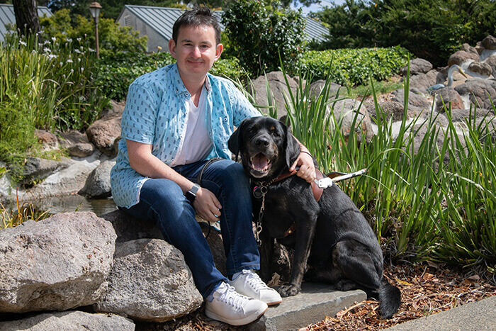 A man wraps arm around black Lab guide dog in a harness and smiles.